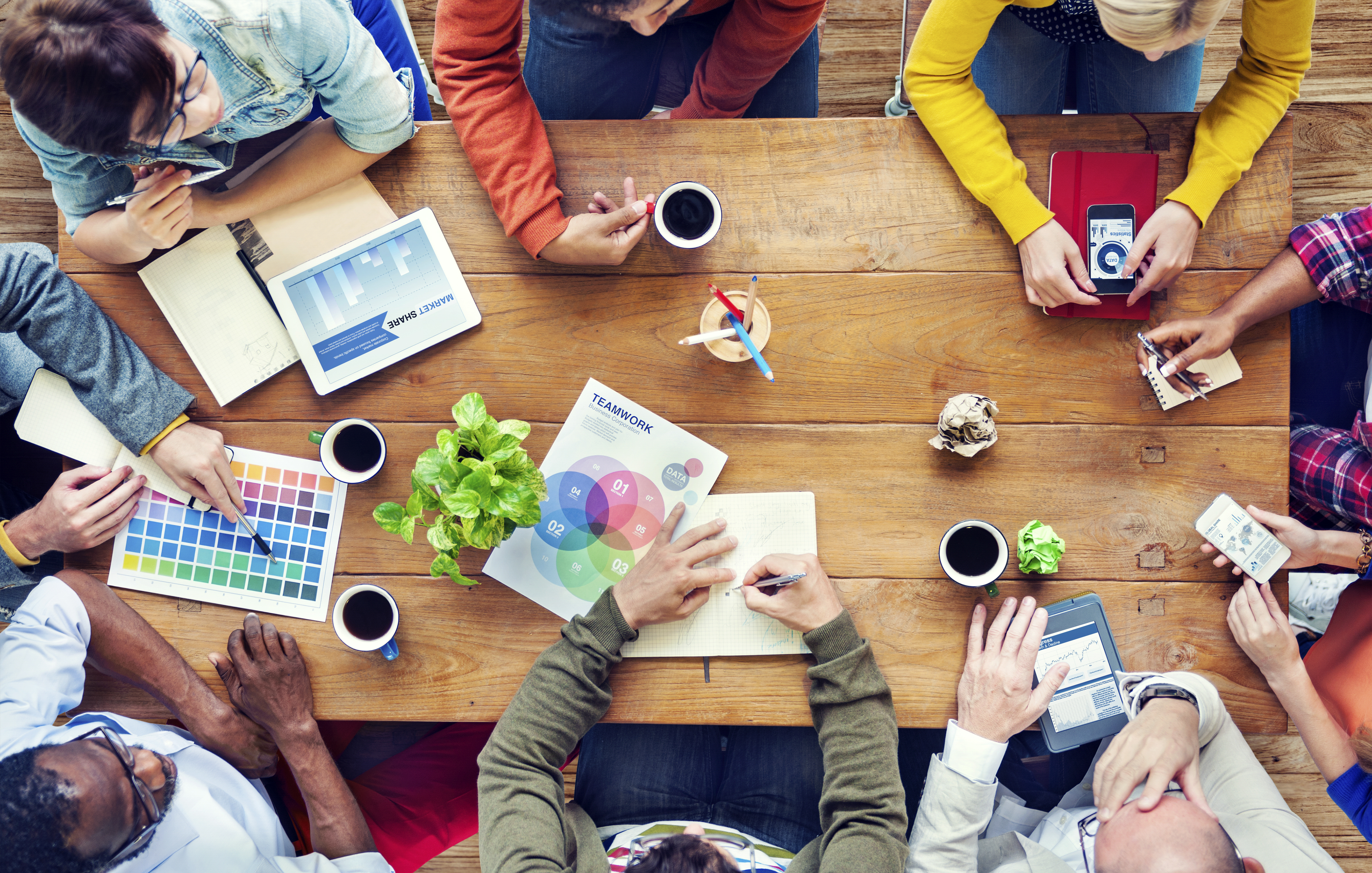 Overhead view of a diverse team collaborating around a wooden table with tablets, charts, color swatches, coffee cups, and a teamwork diagram during a branding and strategy meeting.